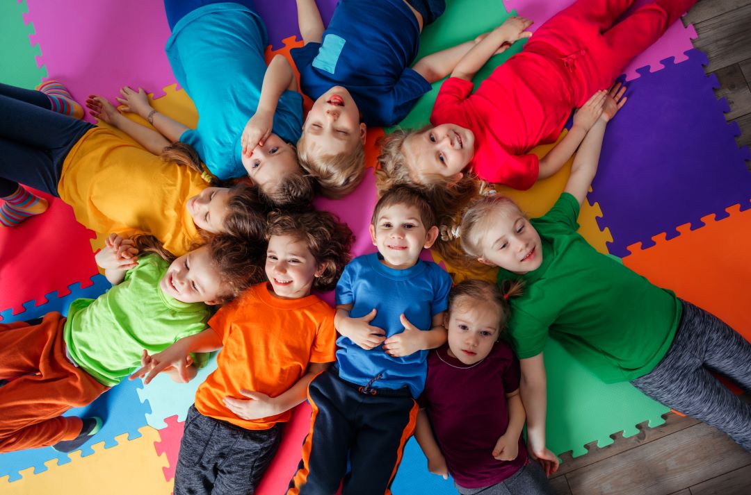 neuf enfants souriants, couchés au sol, avec tee shirt multicolore, sol coloré aussi, photo prise de haut.